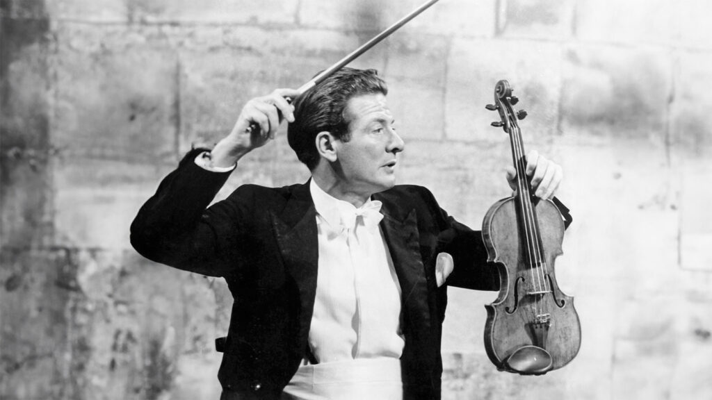 Against the historic stone backdrop of Dartington Great Hall: Neville Marriner with violin in left hand makes an effort to conduct from the front desk with his bow.